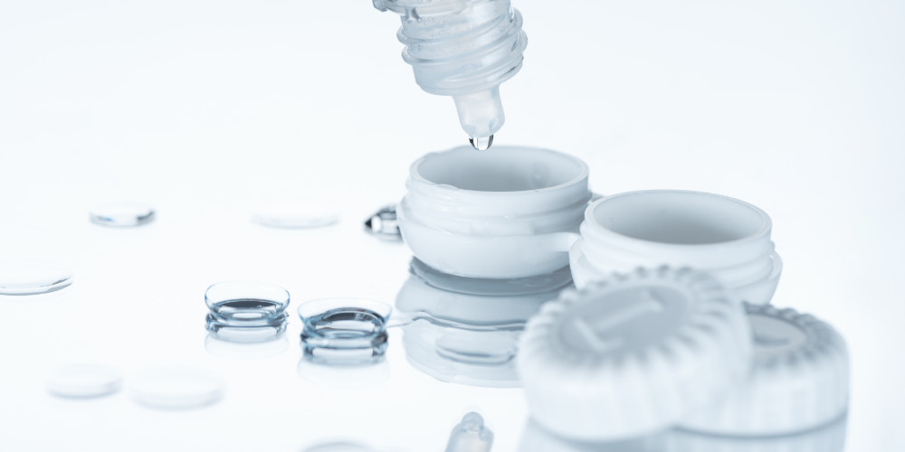 Ein Tropfen treufelt aus einer Plastikflasche in ein Kontaktlinsen Behälter und daneben liegen die Kontaktlinsen